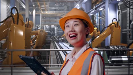 close up side view of asian female engineer with safety helmet looking at the tablet in her hand and looking around while standing with industrial robotic arms working