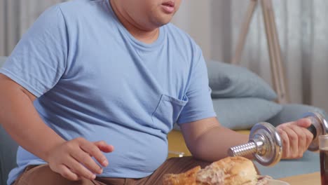 close up of a fat asian man exercising with dumbbell and dipping french fries in ketchup eating fast food on a sofa in the living room at home