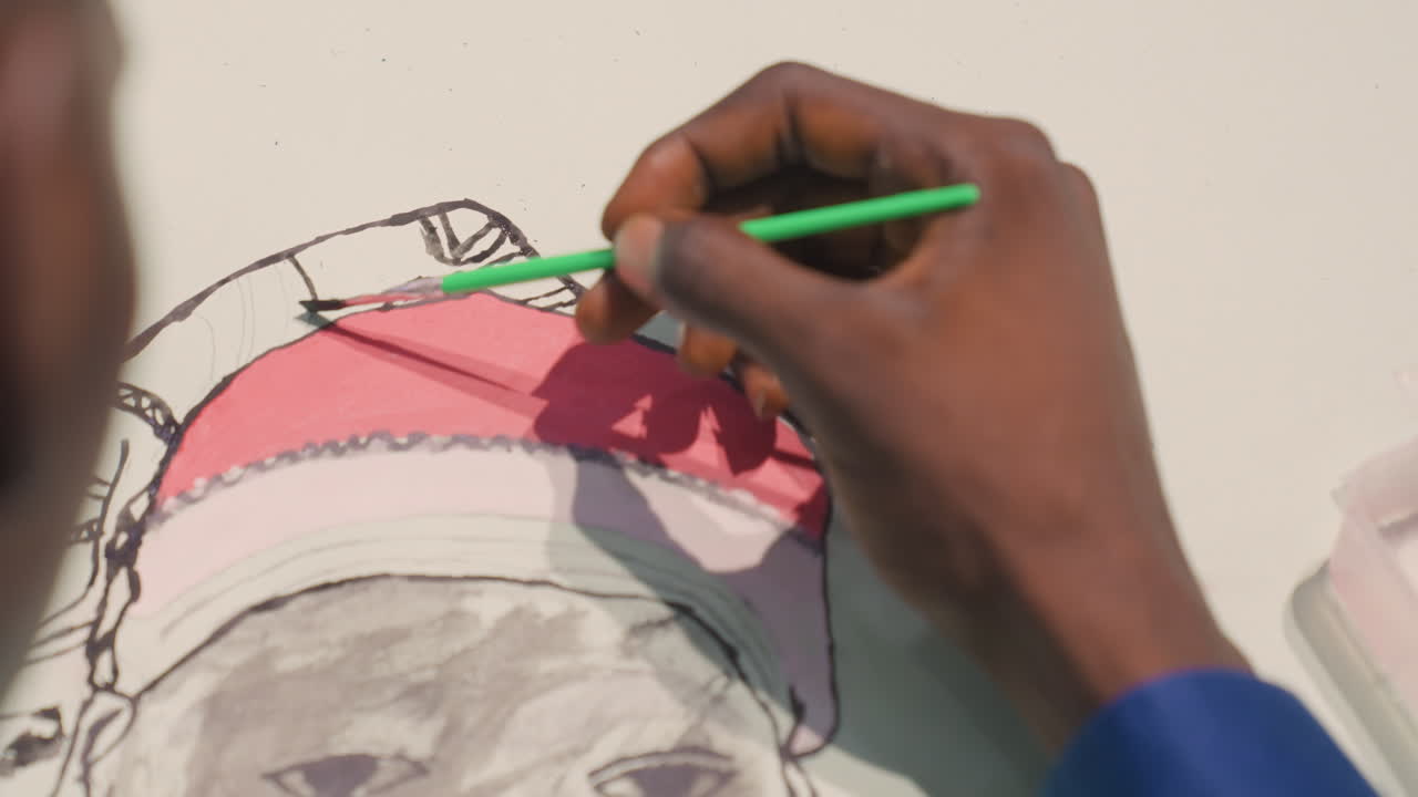 Black illustrator painting hat detail with green brush and steady hand, closeup of pink pigment filling outlined hat in sketch blue sleeve and paper edge visible, deliberate strokes and focused