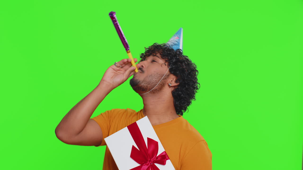 Uomo indiano felice e fortunato che indossa un cappello a cono da compleanno e soffia una trombettina da festa, celebrando il successo e la vittoria