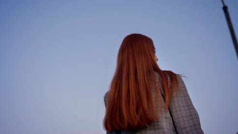 young beautiful woman with long ginger hair looking in camera and turning around, blue sky above, circling around view, low angle