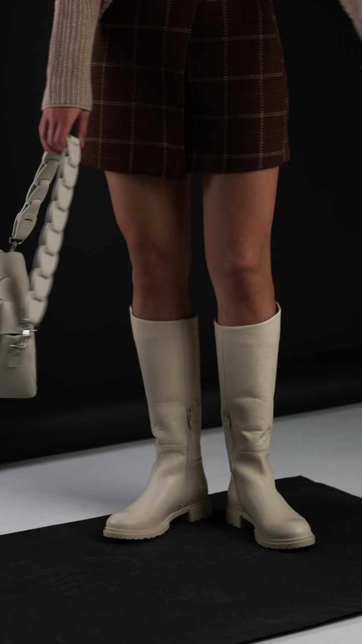 Female demonstrating milk-white high boots on plain soles and same-shade bag. Model turns to show the footwear from different sides. Studio backdrop. Vertical video