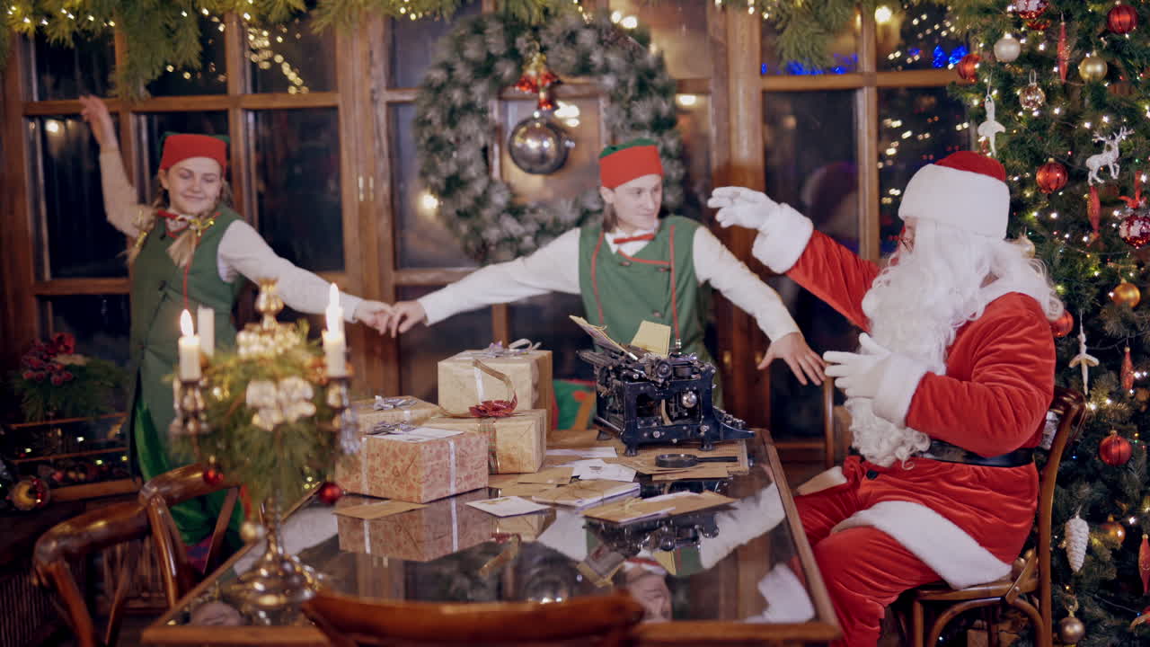 Two merry elves in costumes are dancing together and Santa shows them movements by his hands. Santa Claus is sitting at the table with many gifts and letters on the background of elves.