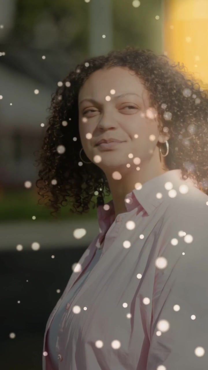Video verticale: Ripresa all'indietro che mostra una donna che guarda il sole vicino a un palo dell'autobus mentre delle particelle le fluttuano intorno.