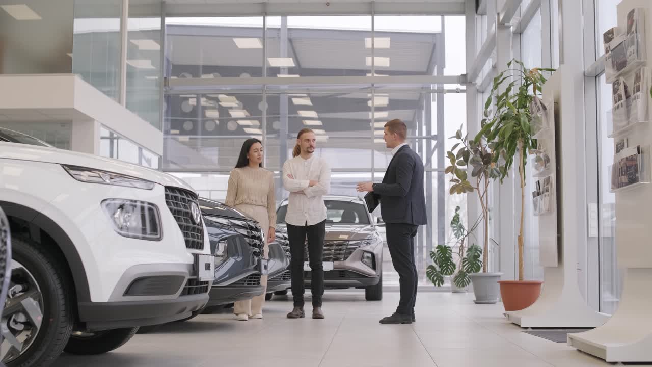 un joven vendedor de coches mostrando a una pareja de jóvenes un nuevo automóvil en el salón de concesionarios.