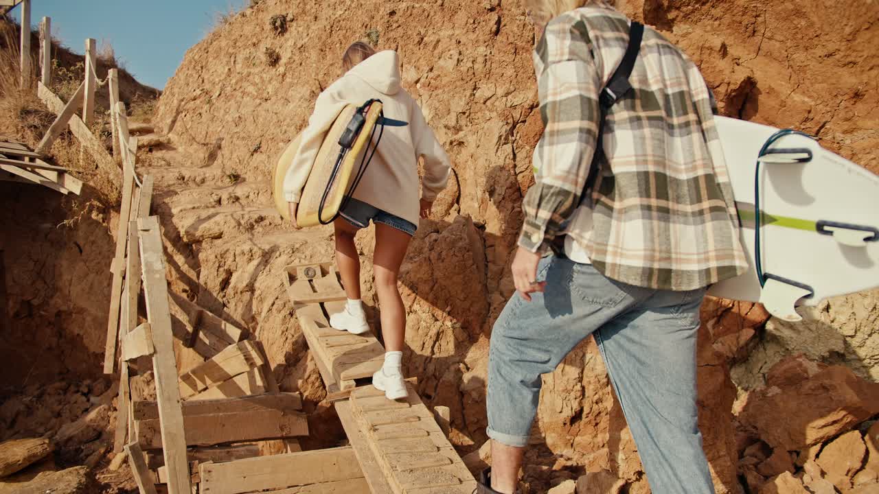 un chico rubio con una camisa a cuadros y su novia rubia con una sudadera blanca suben una escalera de madera en una playa rocosa y llevan tablas de surf con ellos