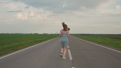 excited woman in denim dress runs joyfully along straight asphalt road marked by white line holding colourful pinwheel high under cloudy sky hair blowing in summer breeze