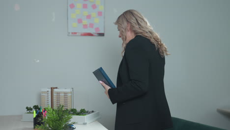 confident businesswoman with wavy blonde hair in black blazer on phone call while holding blue notebook, preparing to sit at desk in modern office environment with shelves in background