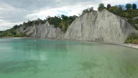 Drone-shot-starting-over-clear-water-at-Scarborough-Bluffs,-flying-up-towards-trees-with-green-and-orange-leaves-revealing-housing-and-buildings-in-the-city-of-Scarborough,-Ontario,-Canada