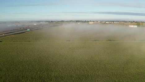 light covering of fog hanging over farm field in late morning seen from drone flying toward town on horizon