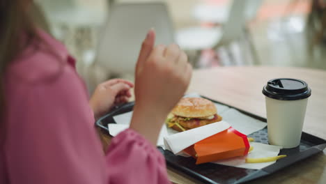 mujer de vestido rosa vista por detrás, disfrutando de papas fritas, hamburguesa y café en una bandeja negra, recoge una patata frita, la come y sacude la cabeza, saboreando el sabor, mientras está sentada en una mesa de madera