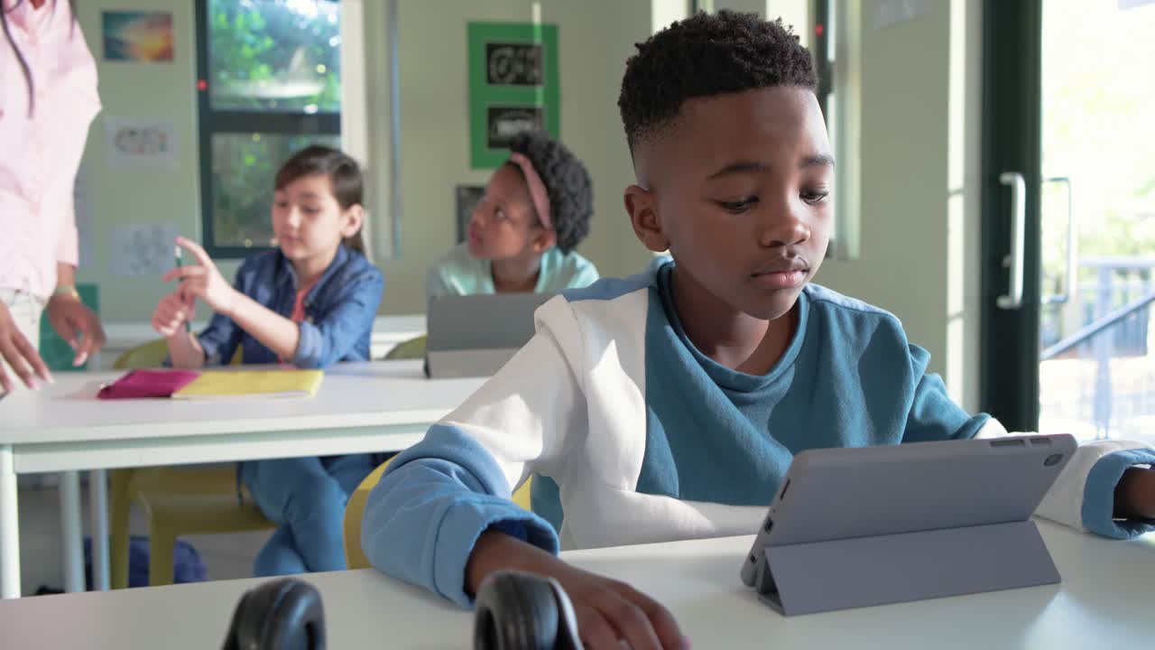 Un ragazzo afroamericano usa un tablet alla scrivania, mentre l'insegnante entra da sinistra e lo assiste sorridendo con un foglio giallo.