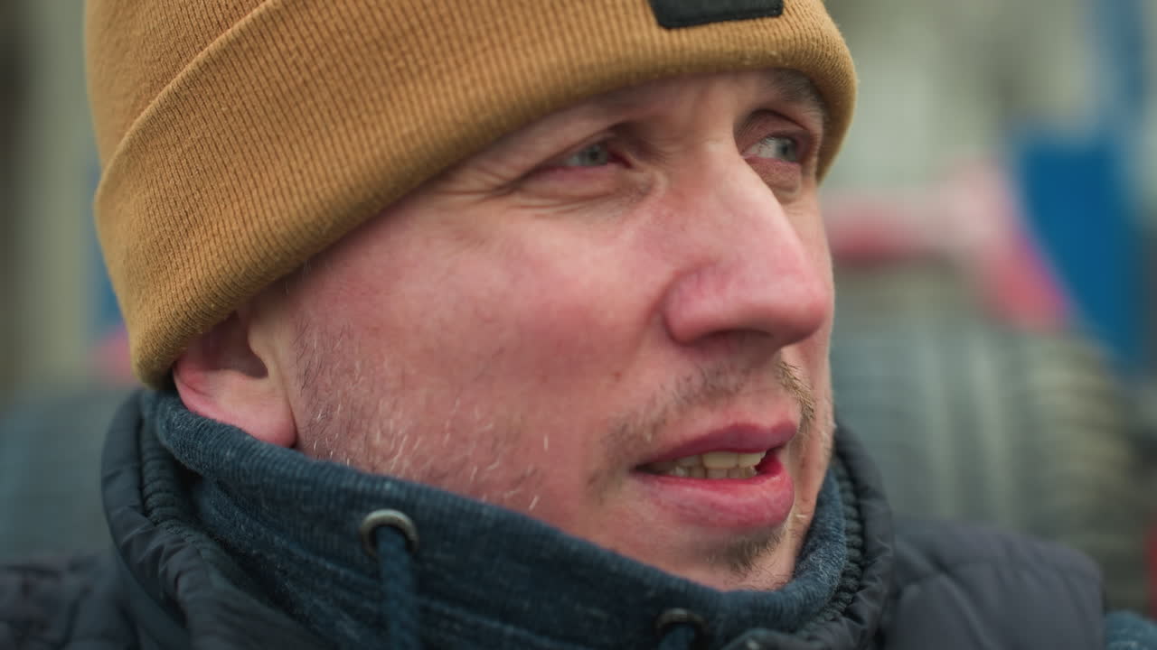 Close-up of a man wearing a brown beanie and black jacket, looking focused into the distance, the background is blurred