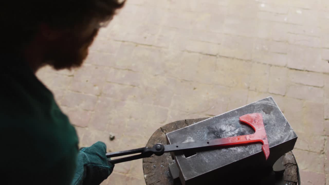 Caucasian male blacksmith wearing safety glasses, hammering hot metal tool on anvil in workshop