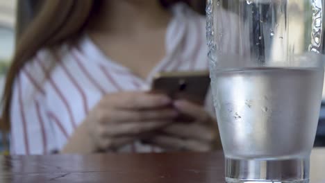 chica fuera de foco jugando con su teléfono inteligente detrás de un vaso de agua enfocado