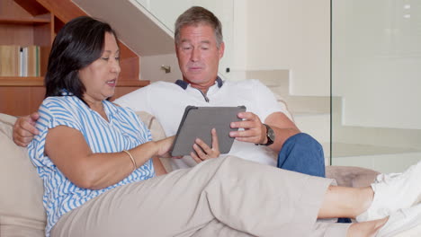 senior diverse couple relaxing on sofa, using tablet and discussing content together