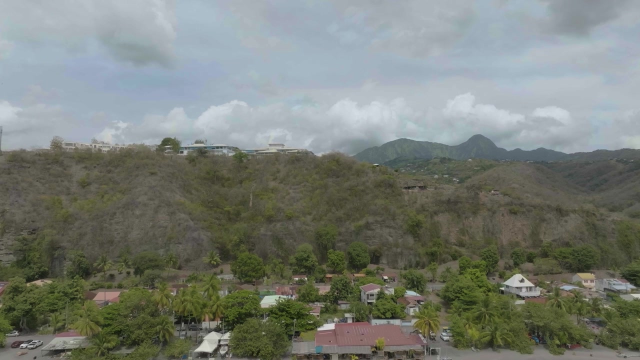 Ripresa statica da drone che mostra dall'alto la piazza principale del quartiere in Martinica, Francia, dove si vede un ristorante effet mer, l'ospedale principale centre hospitalier nord caraibe e il vulcano Pelée nell'entroterra, aperto a modifiche