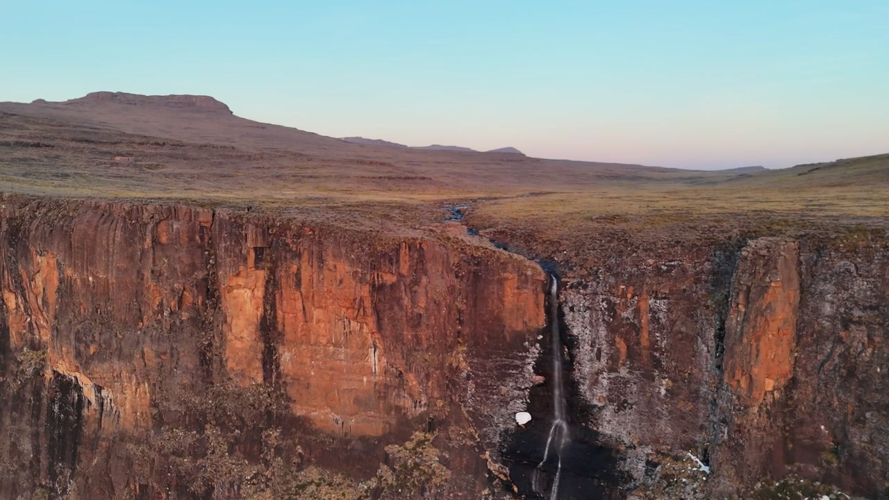 Il drone avanza verso una sottile cascata che scende a cascata su una scarpata verticale di roccia basaltica al tramonto su un altopiano montano