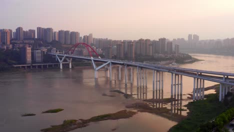 Hazey-yellow-glow-in-sunset-sky-above-urban-Chongqing-city-china-large-highway-bridge-spanning-river,-aerial-panoramic