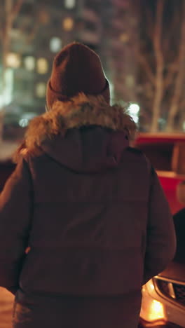 vista en primera persona de una mujer con un abrigo de invierno con una capucha de piel, caminando hacia un coche negro por la noche en un estacionamiento. el fondo presenta coches estacionados y edificios iluminados