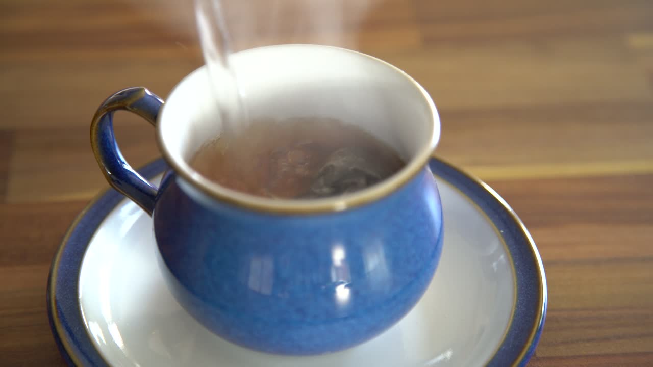 el agua caliente se enroca sobre una bolsita de té que da como resultado un té marrón humeante en una taza de té azul sobre una mesa de madera