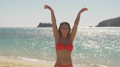 mujer caucásica sexy en bikini rojo de pie en la playa, levantando los brazos bajo la luz del sol