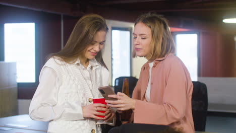 two female colleagues holding drinks and looking something on the mobile phone during a party in the office