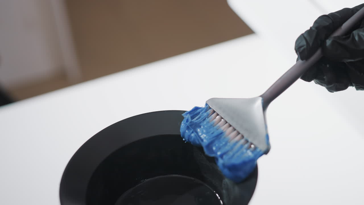 Primer plano de una asistente que saca tinte azul de un tarro con un pincel, textura de pasta espesa en el aplicador, mostrador blanco limpio y estación de mezcla, paso de preparación deliberado antes de la aplicación en el salón.