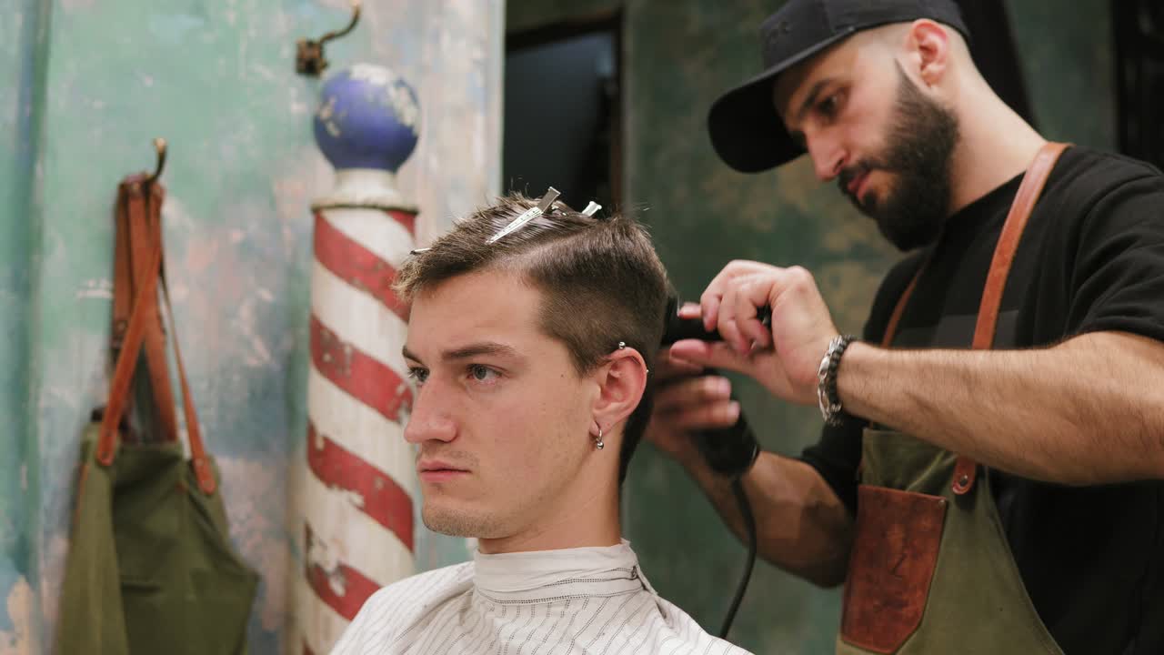 un joven hipster serio siendo cortado el pelo por un barbero profesional en un tiro de barbero. un joven barbero barbudo de pie y haciendo elegante