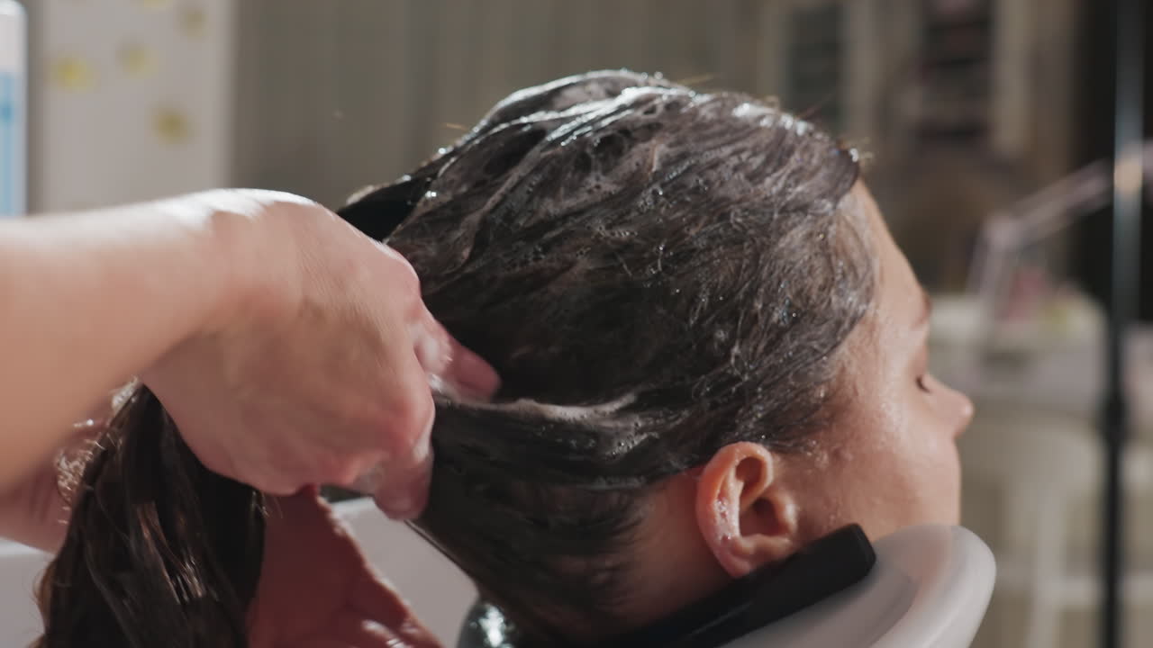 Donna caucasica distesa mentre uno stilista esegue un massaggio profondo e risciacquo del cuoio capelluto con schiuma di shampoo, schiuma lussuosa, trattamento tattile rilassante, ambiente luminoso e professionale del salone e cura post-trattamento calmante