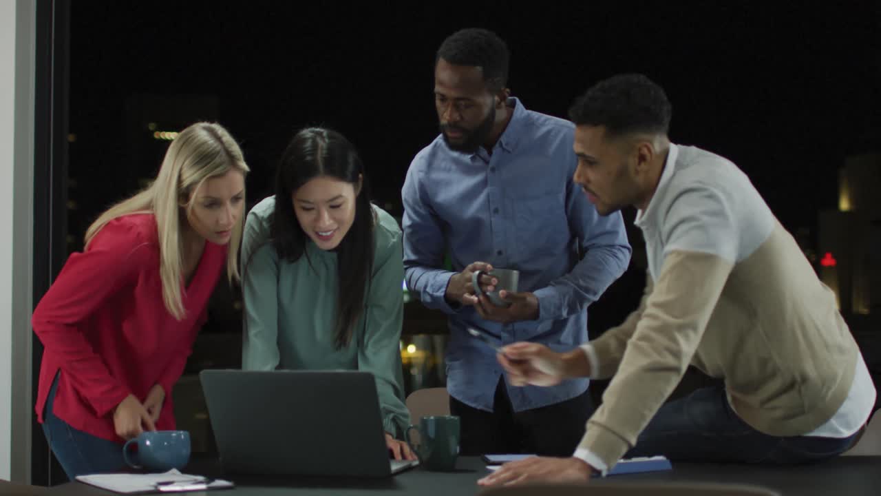 Video of diverse group of colleagues talking standing around a laptop at night in office