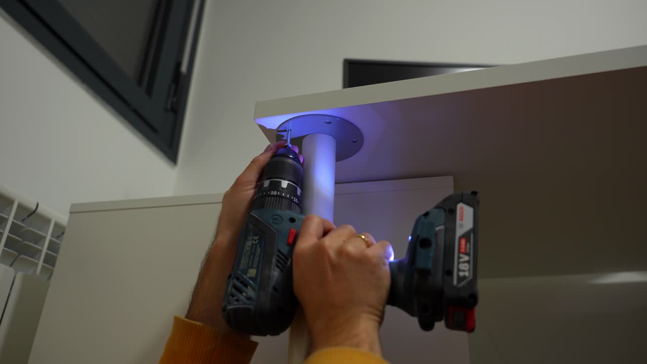 Video footage of man using electric screwdriver to fix metal leg on wooden table at home
