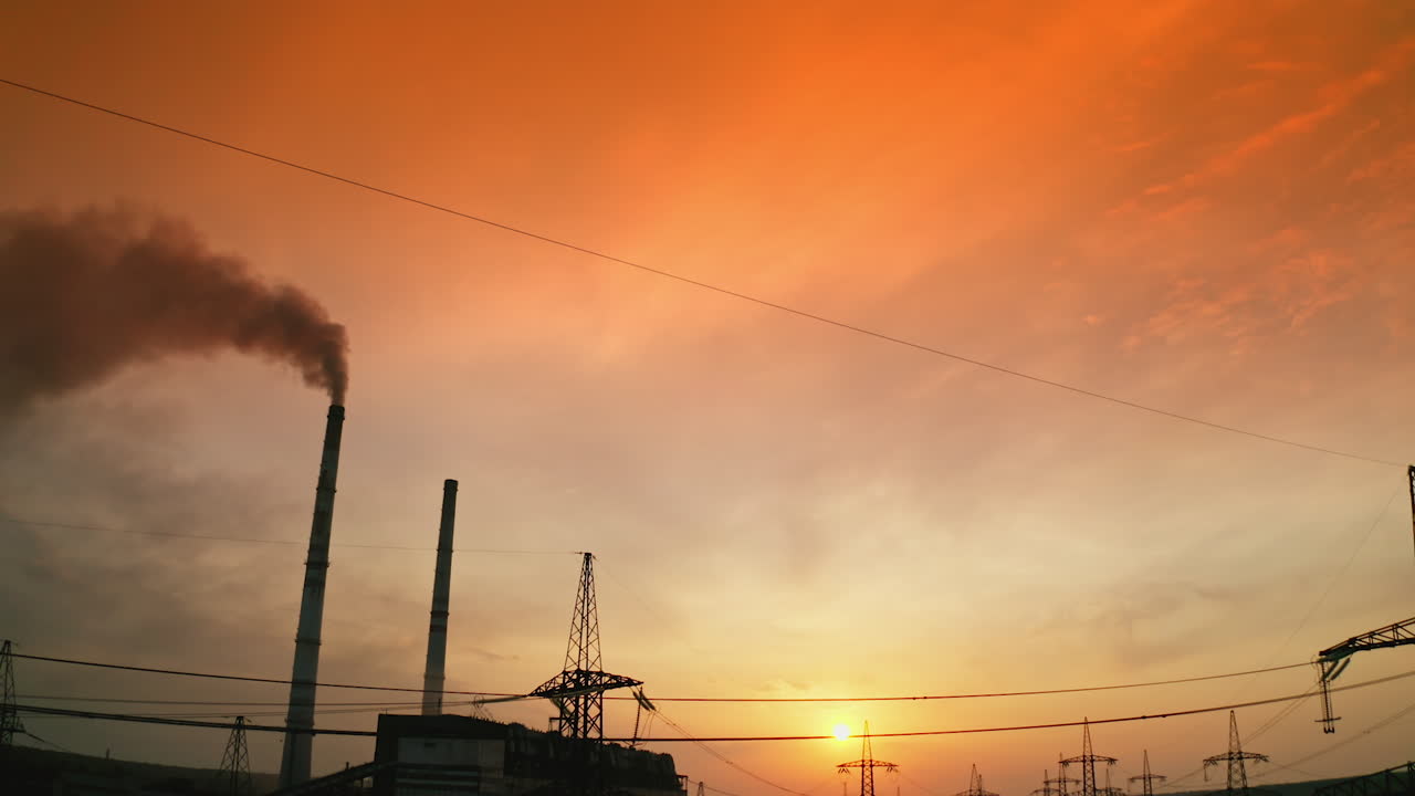 Pipe with dark smoke near the transmission lines. High-voltage power towers next to the harmful manufacture against the epic sky of setting sun. Motion top down.