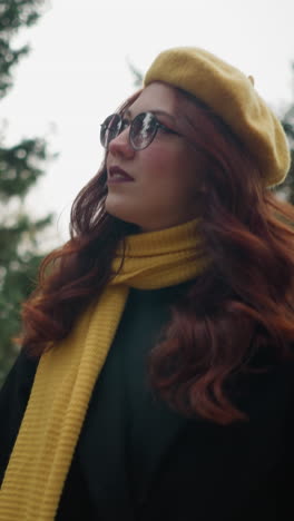 una dama universitaria de cabello rojo fluido mira hacia arriba, vestida con un abrigo negro, boina amarilla y una bufanda a juego, vagando por un exuberante parque adornado con colores de otoño