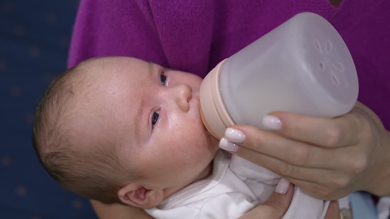 Beautiful newborn boy resting in mother's hands and being fed from a bottle. Little child closing eyes and gradually falling asleep. Close up.