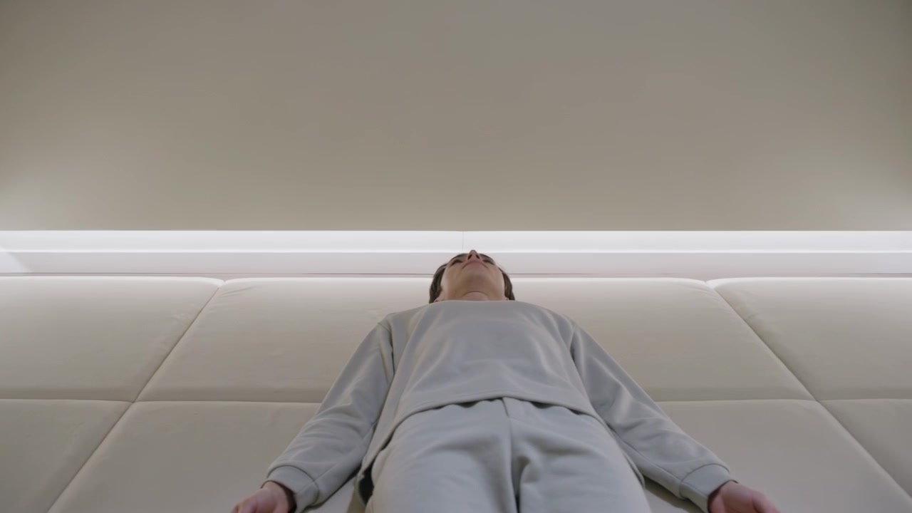Lying Asian man resting, tilting head back as frames progressing under LED light, gray loungewear