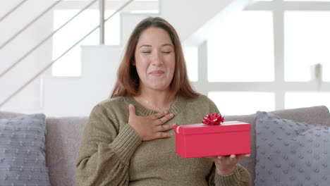 excited plus size biracial woman having video call, opening gift in living room, slow motion