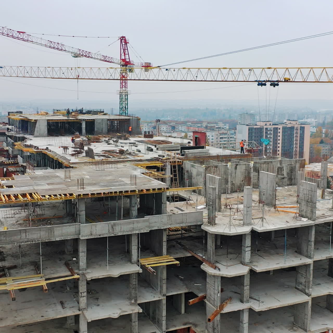 New construction site of shopping center. Aerial view on roof with workers and big cranes during construction on the city background. Drone moves back.