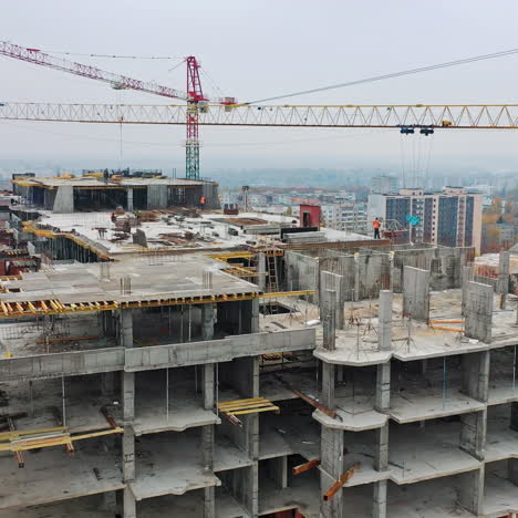 new construction site of shopping center. aerial view on roof with workers and big cranes during construction on the city background. drone moves back.