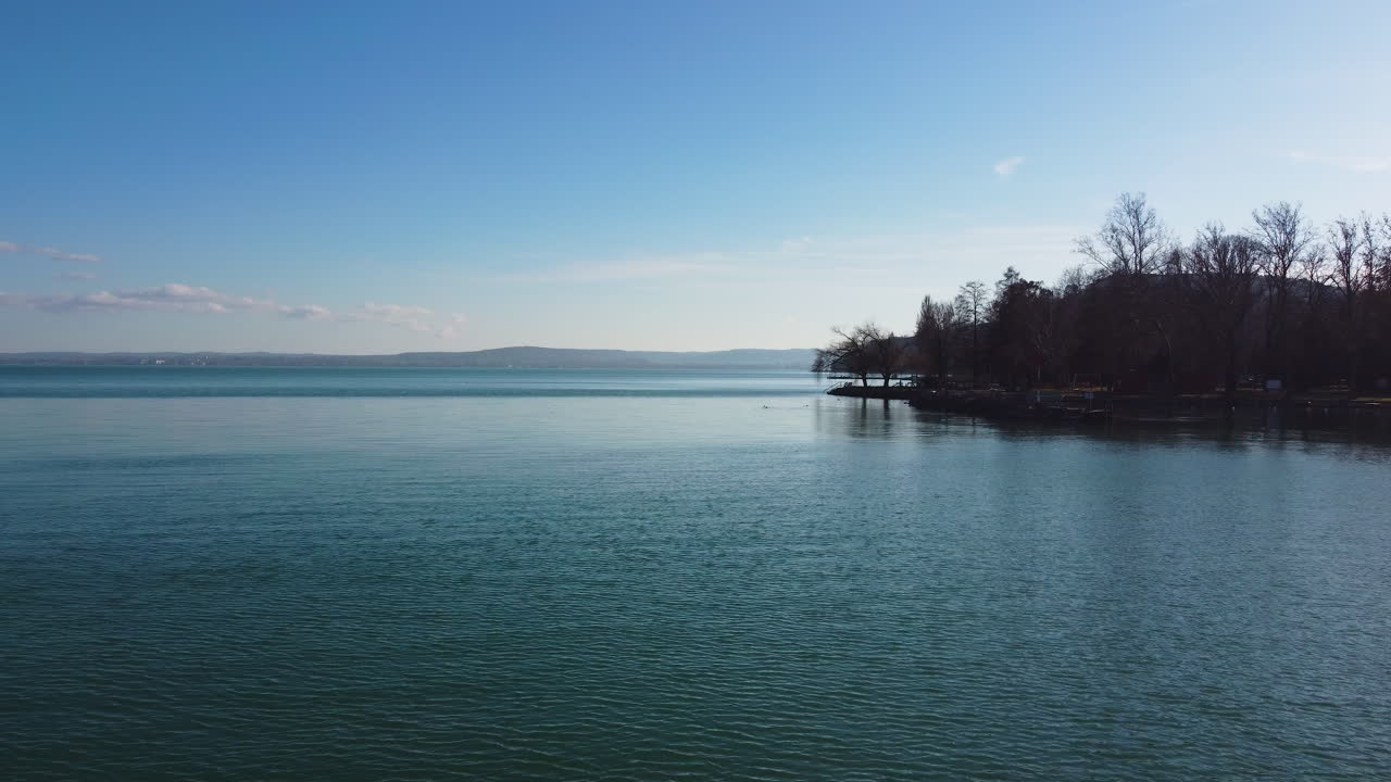 Sunlit lakeshore view with tree silhouettes on Lake Balaton, calm reflective water in foreground