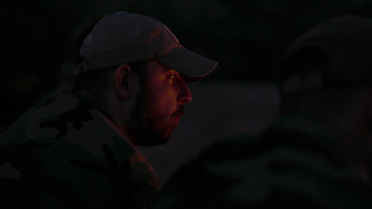Close-up of a bearded man's face in camouflage clothes and cap at night lit by campfire flames, he tells a scary story to his friends, night camp, hunting