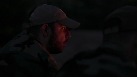 close-up of a bearded man's face in camouflage clothes and cap at night lit by campfire flames, he tells a scary story to his friends, night camp, hunting