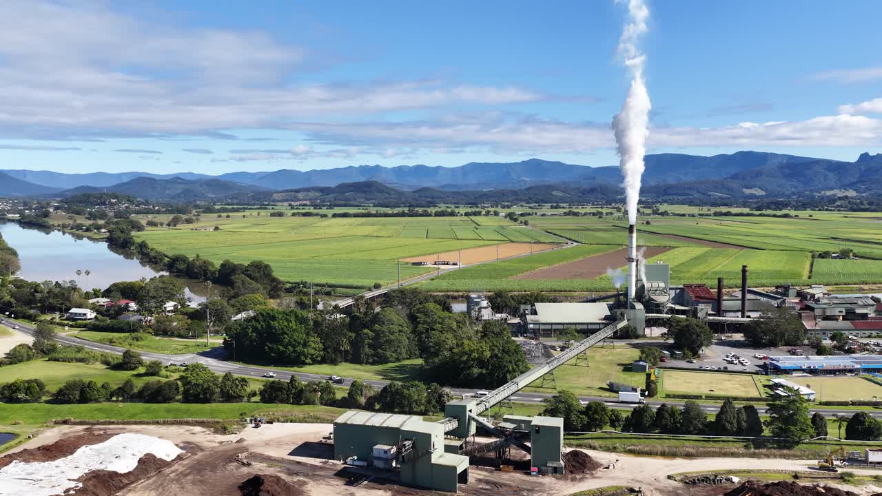 Aerial footage captures a sugar mill with smoke stacks, surrounded by lush green fields and distant mountains under clear skies