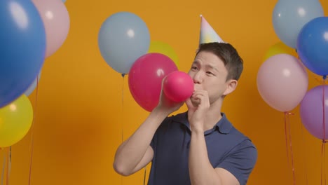 Studioporträt-Eines-Mannes-Mit-Partyhut,-Der-Seinen-Geburtstag-Feiert-Und-Einen-Ballon-In-Die-Luft-Sprengt