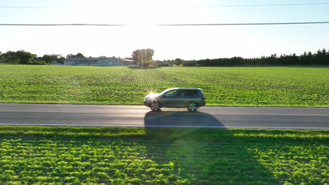 tracking shot of suv traveling along rural road. fields surround car as sunlight reflects off its windshield. travelers cross farmland at sunset