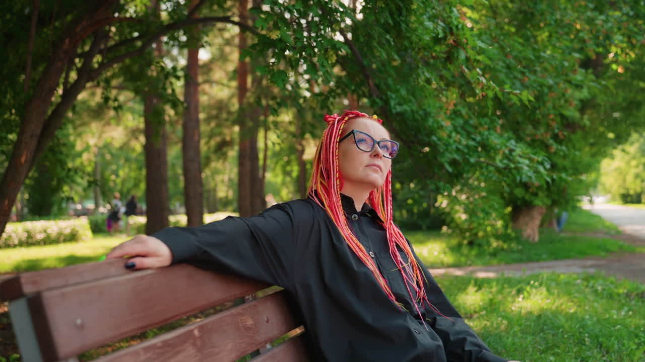artista caucásica con trenzas multicolores recostada en un banco del parque bajo un dosel verde, con las manos apoyadas en el respaldo, los ojos cerrados, soñando despierta, pausa creativa relajada tras dibujar al aire libre, suave moteado