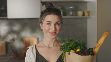 smiling woman holding a bag of fresh groceries in her kitchen