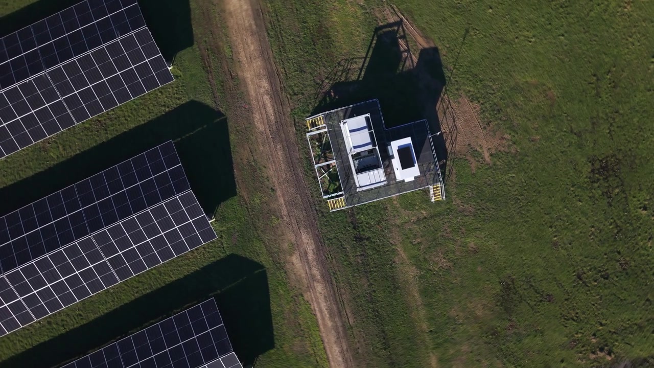 Skarpe droneoptagelser viser rækker af solpaneler og en forsyningsstation i The Basin, Melbourne. Klar sol kaster skarpe skygger på grønt græs, ren energiteknologi og landdistriktsinfrastruktur