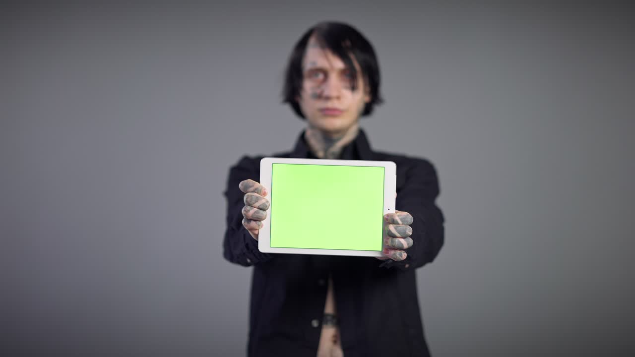 녹색 스크린을 가진 색 태블릿, 회색 배경의 희미한 젊은이와 함께 문신 된 남성 손, 크로마키 기기 포즈를 보여주는 백인 밀레니엄 남자.