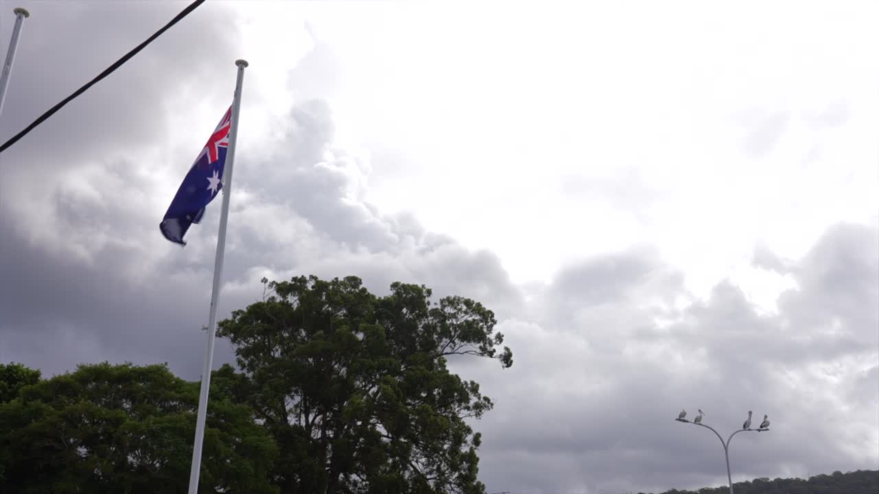 Bandiera australiana che sventola su un pennone con pellicani che osservano, contro un cielo nuvoloso, Woy Woy, Nuovo Galles del Sud, Australia.